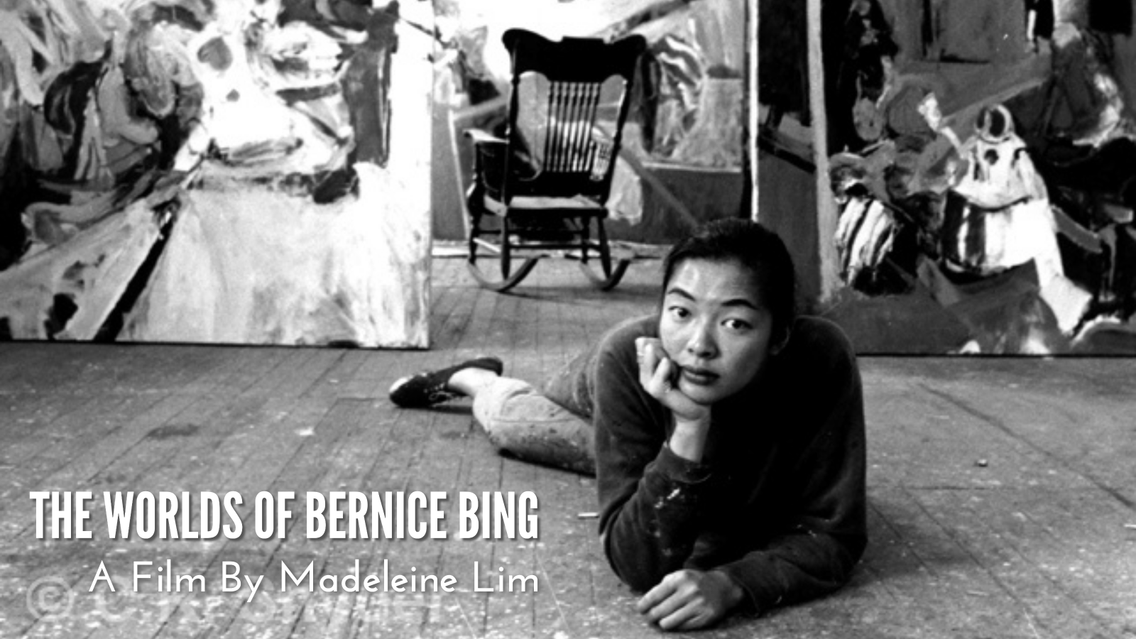 A black and white photo of Bernice Bing, an Asian American woman with black hair, as she lounges on her stomach on the wood-paneled floor and looks into the camera. Behind her, a large rocking chair sits empty between large abstract paintings that lean against the wall.